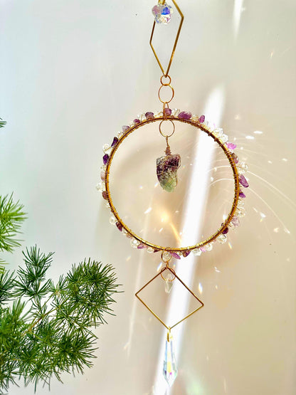 Handmade amethyst and quartz crystal suncatcher hanging in sunlight with indoor plants nearby, casting rainbow reflections on the white wall and showcasing the shimmering raw stone.