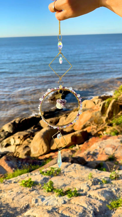 Handmade crystal suncatcher featuring a raw amethyst stone in a golden hoop, accented with faceted beads and geometric brass shapes, held against the ocean background in golden sunlight.