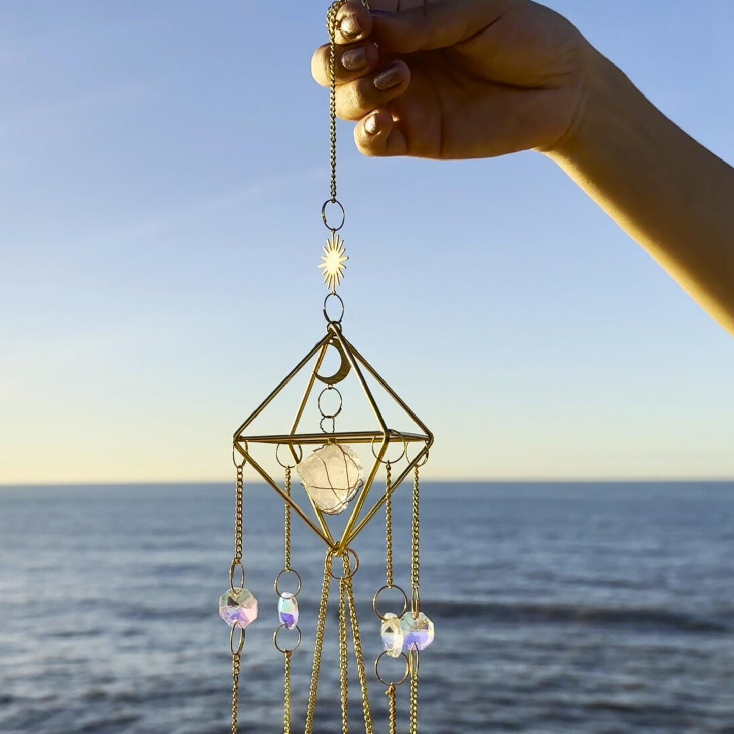 Detail of hanging chains and rose quartz gemstone suspended in a gold
geometric frame, held by hand at the seashore.