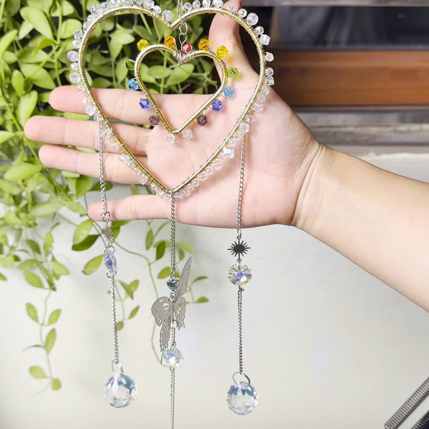 Hand holding a heart-shaped rainbow suncatcher in front of a green plant backdrop. The design features two heart frames wrapped in crystal beads and rainbow colors, with a dangling butterfly and crystal balls.