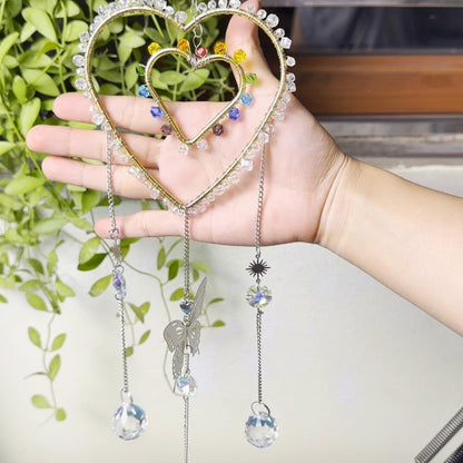 Hand holding a heart-shaped rainbow suncatcher in front of a green plant backdrop. The design features two heart frames wrapped in crystal beads and rainbow colors, with a dangling butterfly and crystal balls.