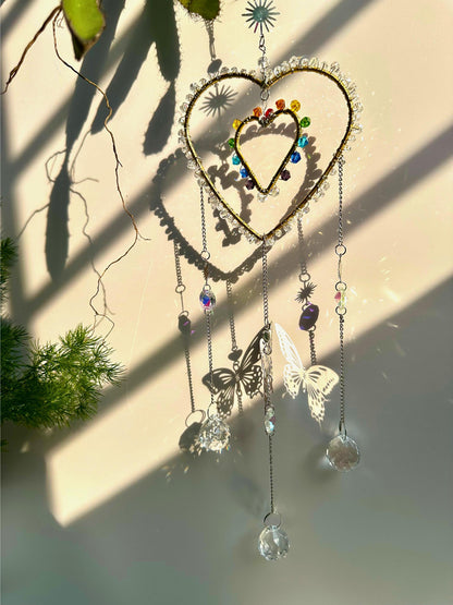 Sunlight shining through a heart-shaped rainbow suncatcher, casting beautiful rainbow reflections and shadows of a butterfly on a white wall.
