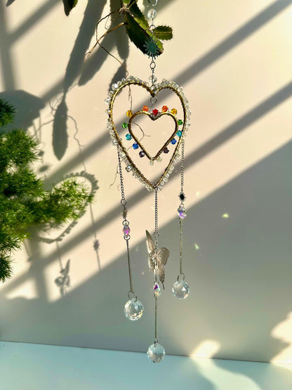 Rainbow crystal heart suncatcher with butterfly charm, suspended near greenery. Three large prism balls hang from chains below, casting dancing rainbow flares.