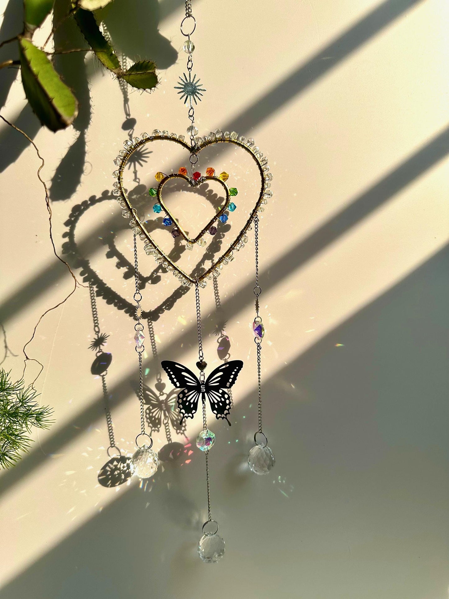 Hanging rainbow heart suncatcher with large butterfly charm in the center, backlit by strong sunlight, projecting vivid crystal reflections and shadows.