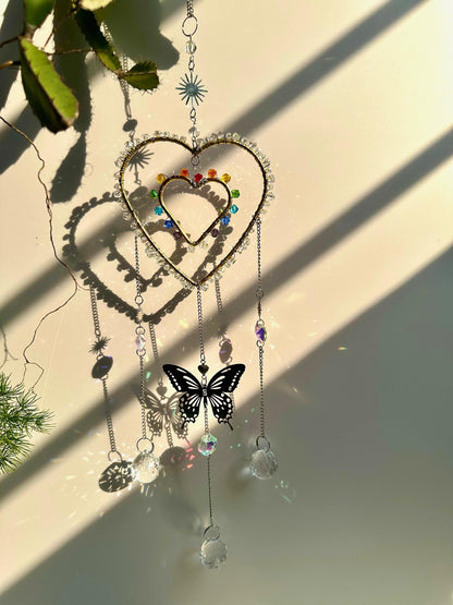 Hanging rainbow heart suncatcher with large butterfly charm in the center, backlit by strong sunlight, projecting vivid crystal reflections and shadows.