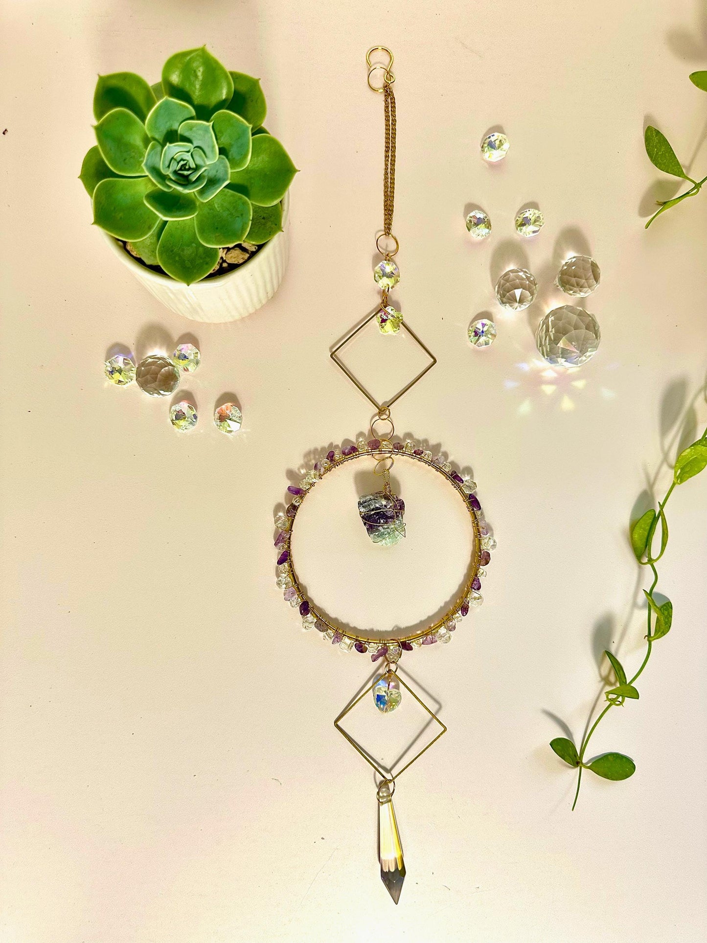 Flat lay of a handmade gemstone and crystal suncatcher displayed with succulents, crystal beads, and greenery on a pastel background, styled for decorative display.