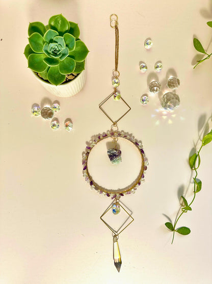 Flat lay of a handmade gemstone and crystal suncatcher displayed with succulents, crystal beads, and greenery on a pastel background, styled for decorative display.