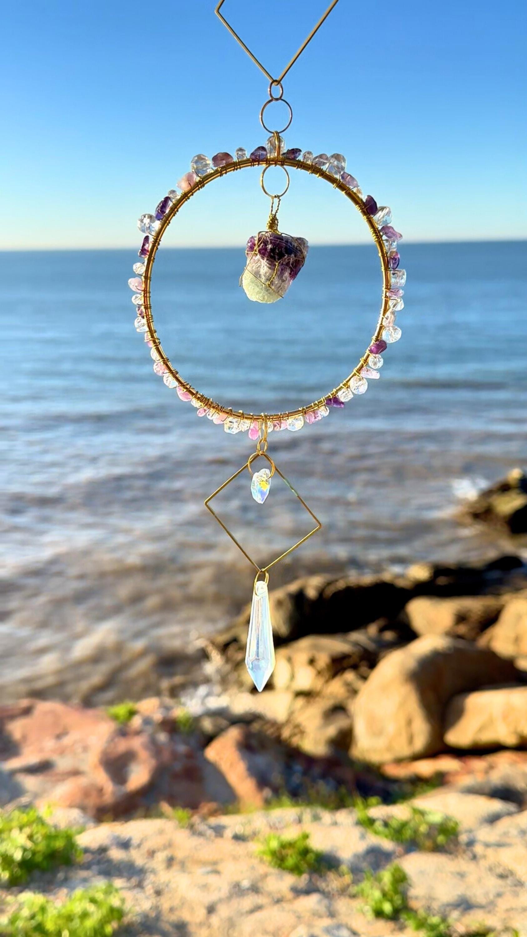 Close-up of a wire-wrapped raw amethyst stone suncatcher, suspended inside a gemstone-adorned gold hoop with crystal pendant, photographed by the sea under clear blue skies.