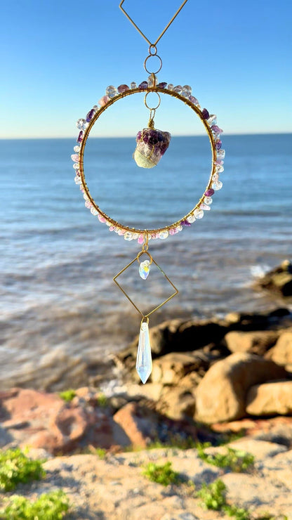 Close-up of a wire-wrapped raw amethyst stone suncatcher, suspended inside a gemstone-adorned gold hoop with crystal pendant, photographed by the sea under clear blue skies.