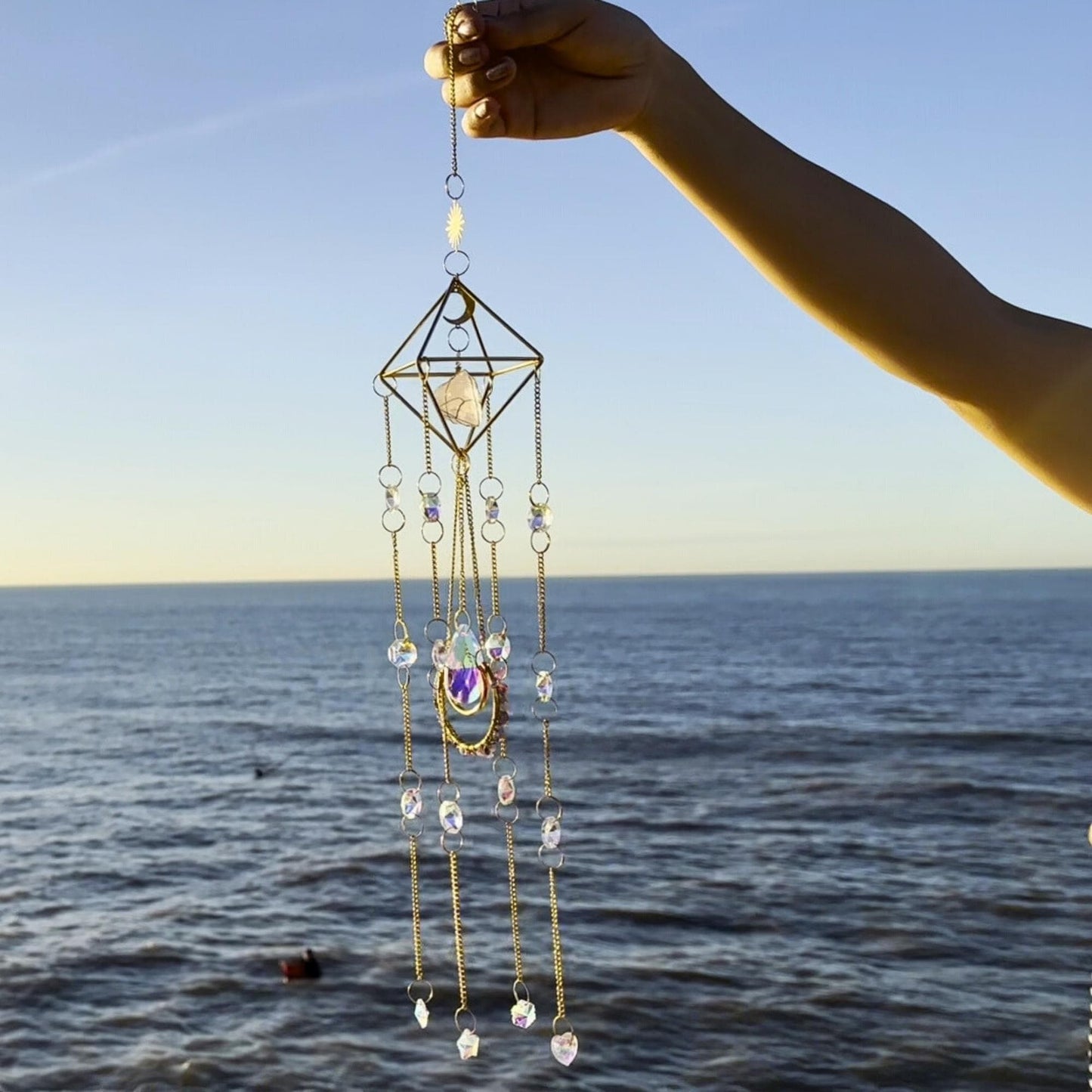 Handmade crystal suncatcher featuring a geometric frame and rose quartz
centerpiece, with cascading strands of iridescent beads and moon-themed
charms, photographed at the beach during golden hour
