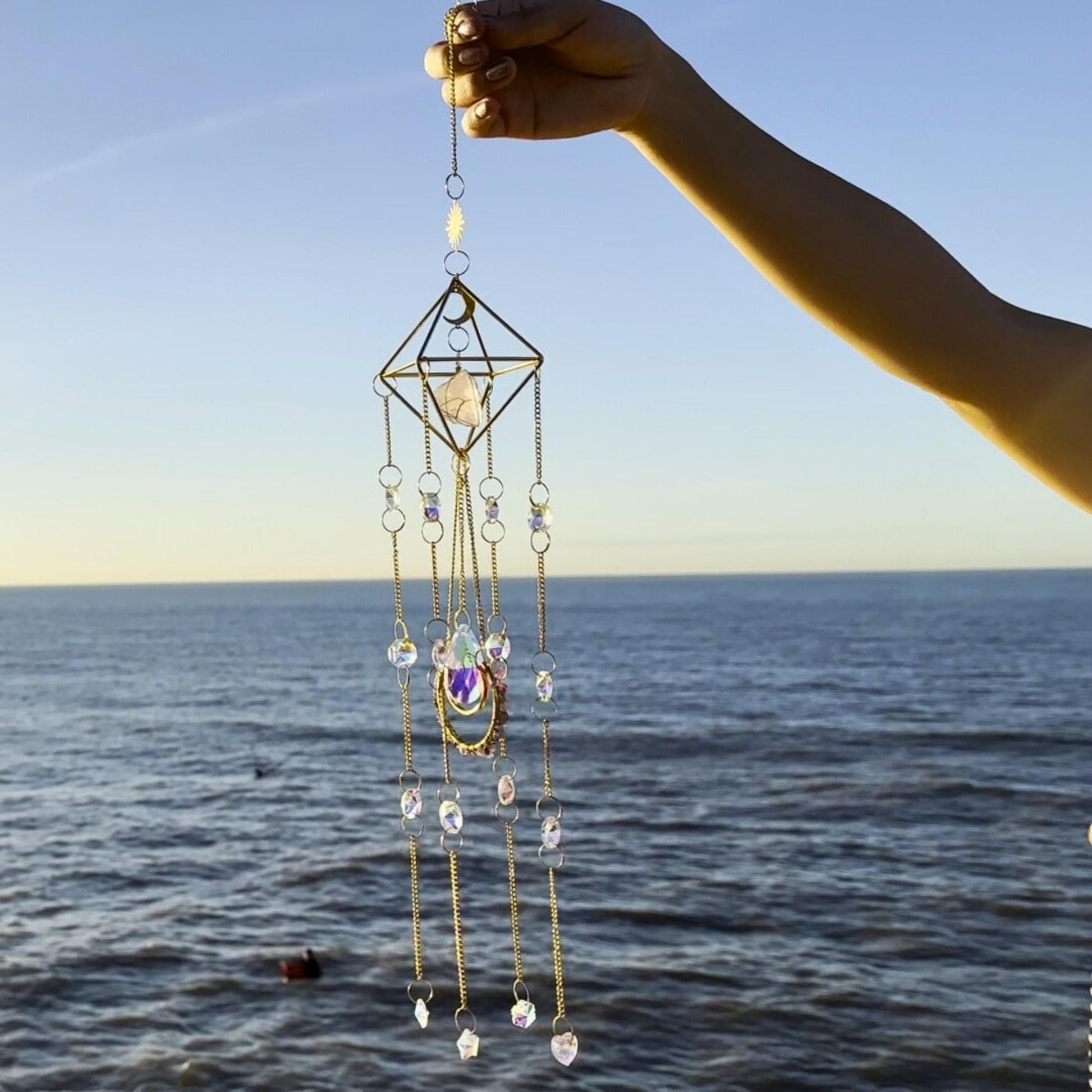 Handmade crystal suncatcher featuring a geometric frame and rose quartz
centerpiece, with cascading strands of iridescent beads and moon-themed
charms, photographed at the beach during golden hour