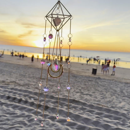 Beach view showcasing a dangling crystal suncatcher with crescent moon
charm and pink-tinted prism centerpiece, glowing under sunset sky