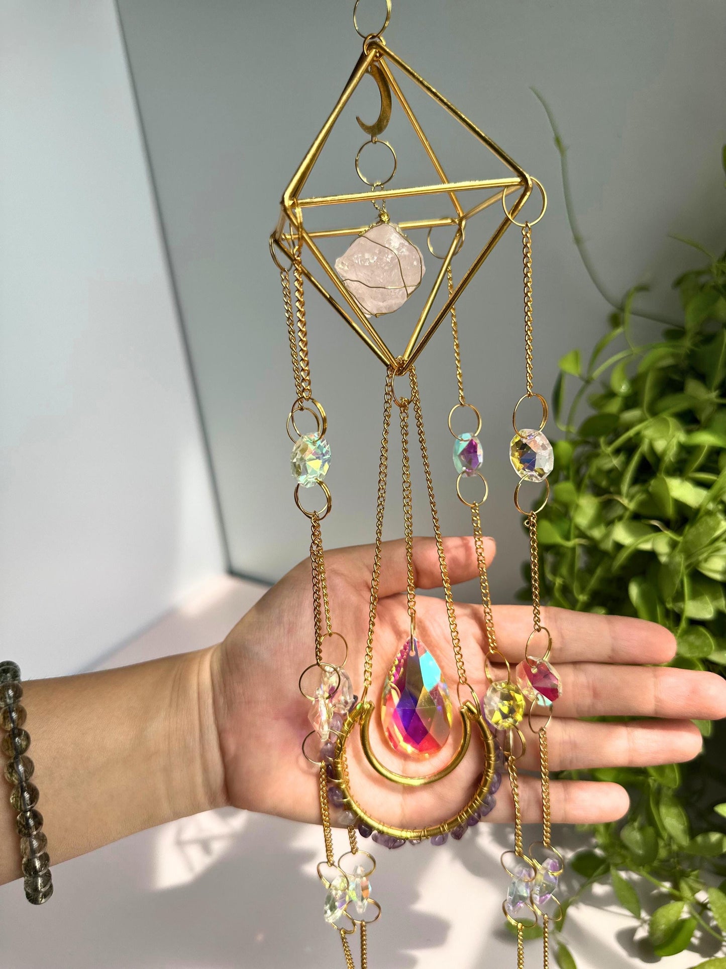 Indoor close-up of a crystal moon catcher with faceted prism, gold chains,
and amethyst accents, held by hand in soft daylight.