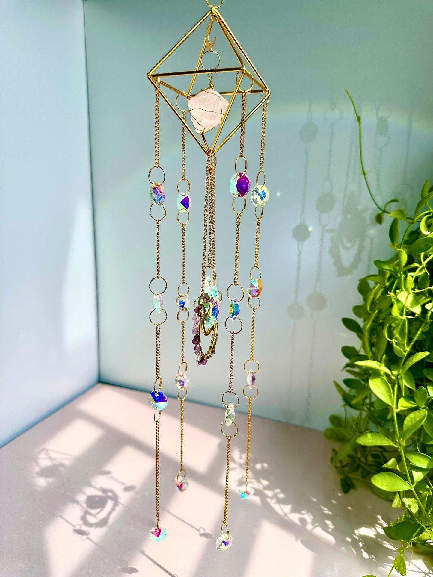 Sunlit crystal mobile with golden geometric frame and rainbow-reflective
beads, displayed indoors with cascading light rays.