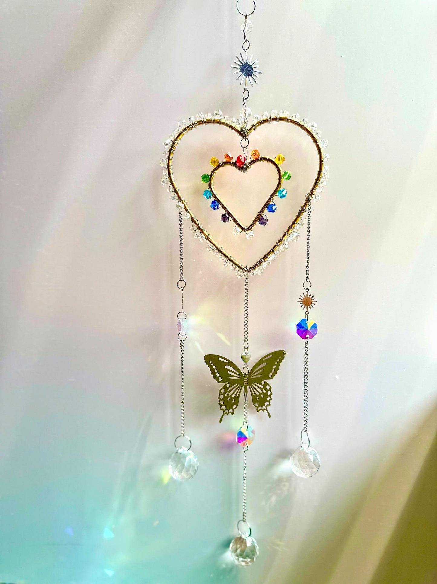 Full view of a rainbow crystal heart suncatcher against a clean pastel background. Featuring a central butterfly charm, crystal strands, and sparkling light refractions below.